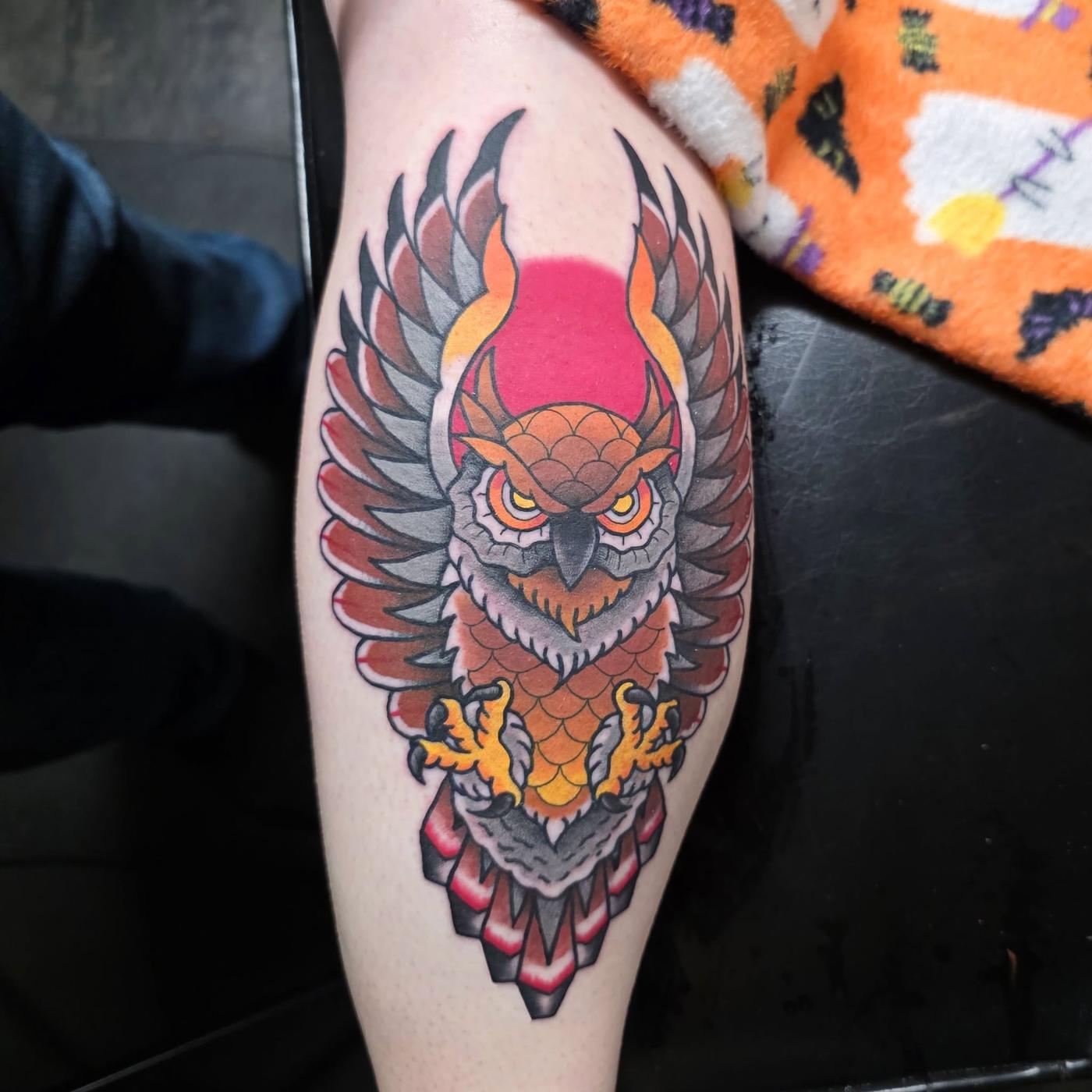 A color traditional owl tattoo on a calf, wings spread over a red shape