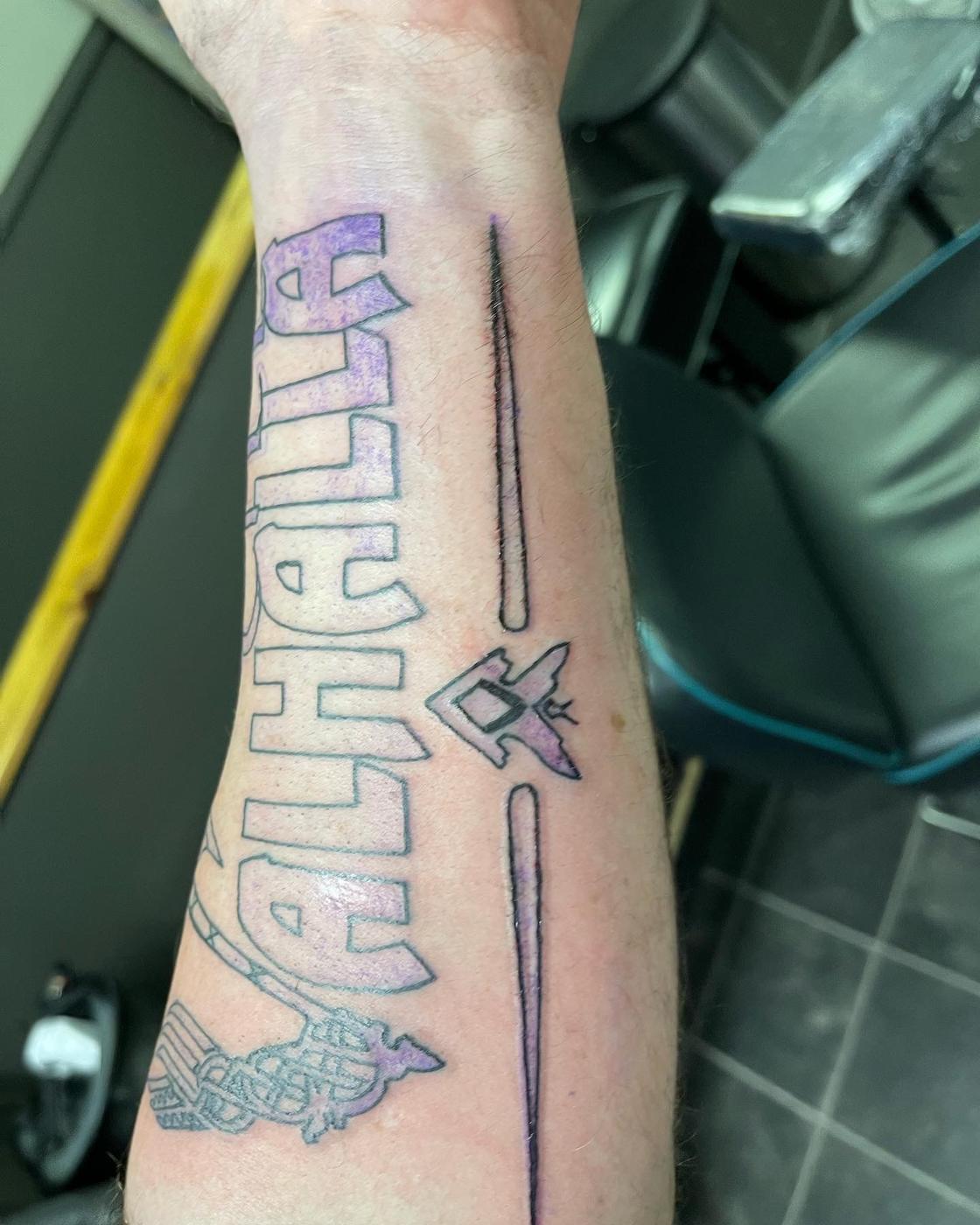 Lettering reading Valhalla with arrows running down a forearm, in color